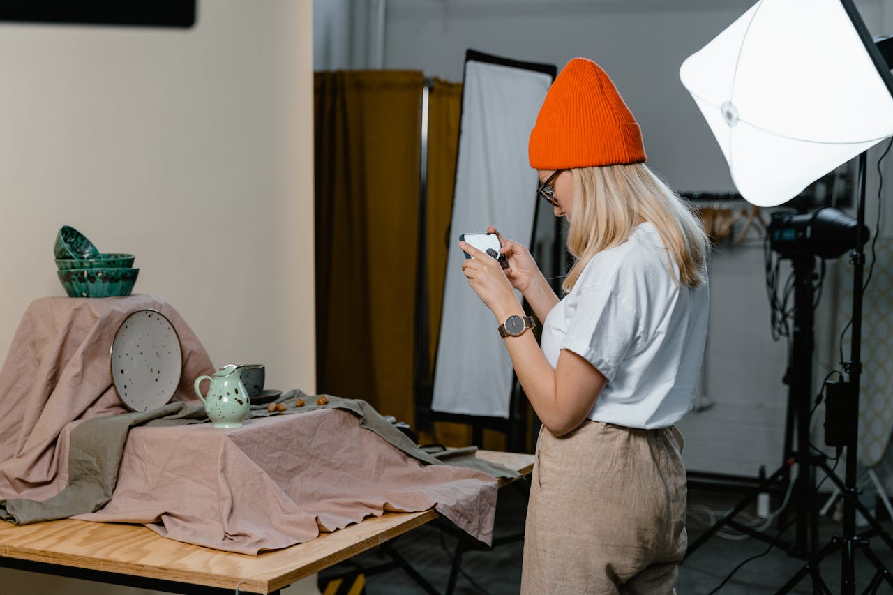 A woman in a beanie takes photos of ceramic items in a studio setup.