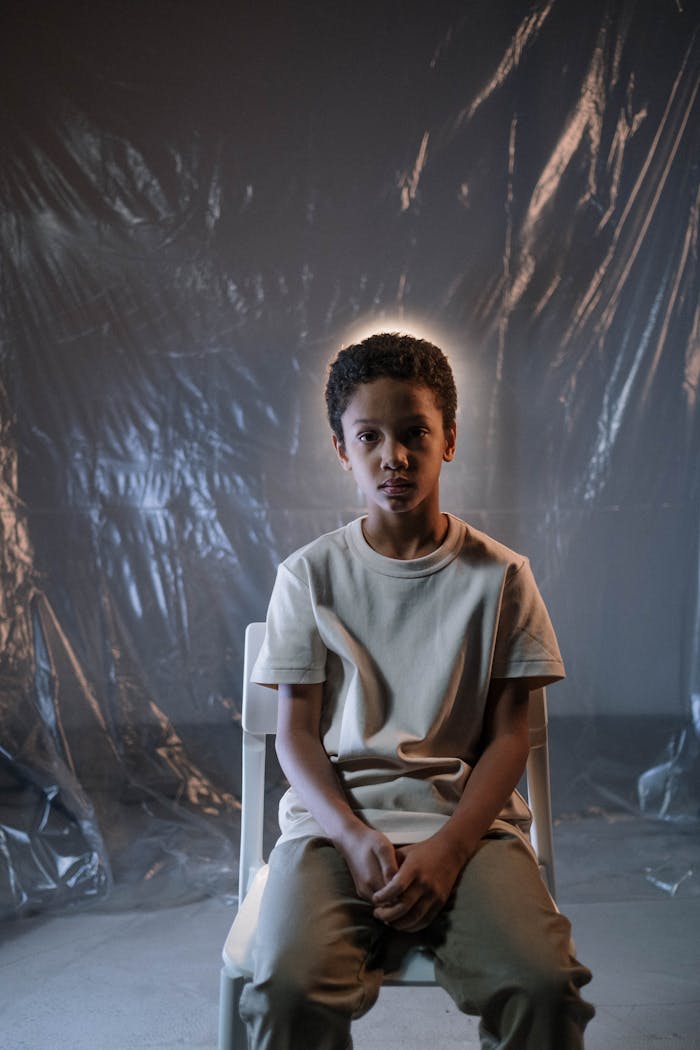 A young boy sitting indoors with dramatic lighting and a plastic backdrop, conveying introspection.